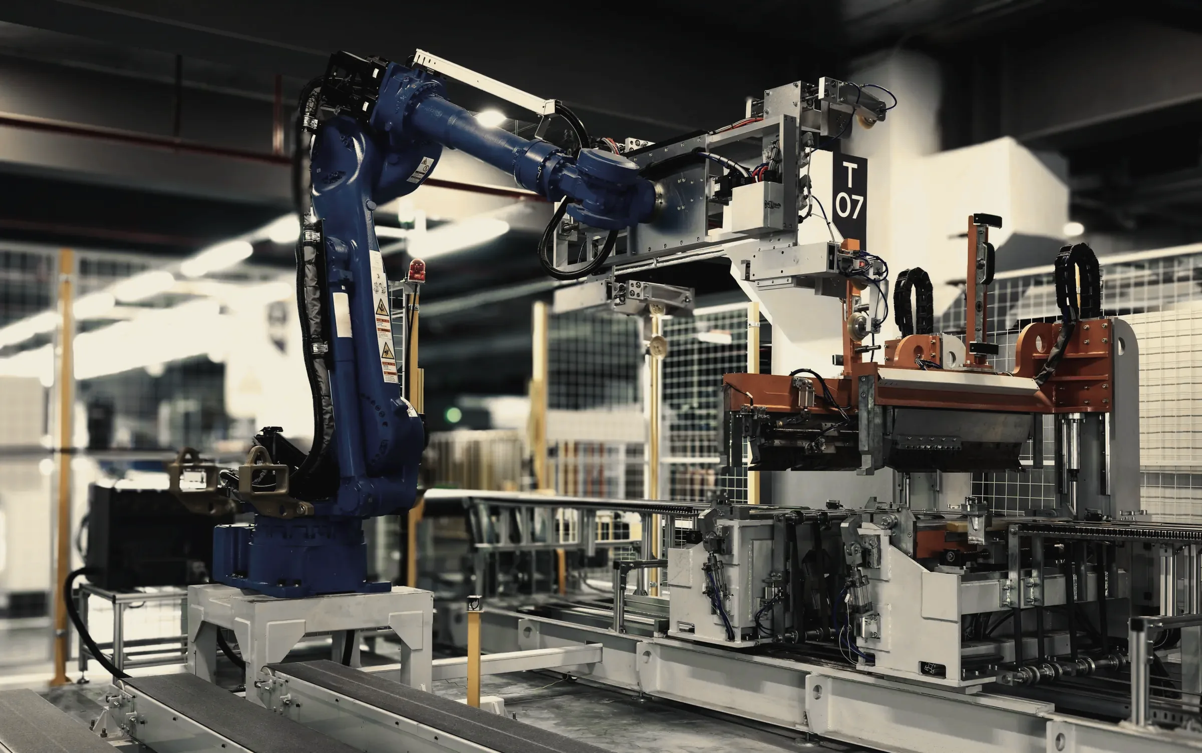 A robot arm in a guarded cell loads parts into a station on a production line.